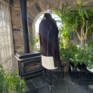 May include: A dark brown suede jacket with cream-colored accents is displayed on a mannequin. The jacket features decorative stitching and a unique design. The background includes a stone wall, plants, and a fireplace, suggesting a rustic or vintage aesthetic.