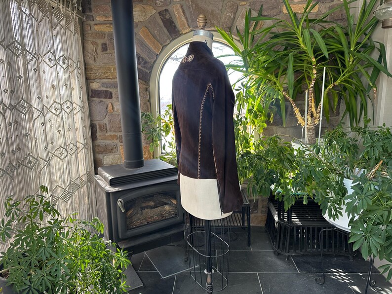 May include: A dark brown suede jacket with cream-colored accents is displayed on a mannequin. The jacket features decorative stitching and a unique design. The background includes a stone wall, plants, and a fireplace, suggesting a rustic or vintage aesthetic.