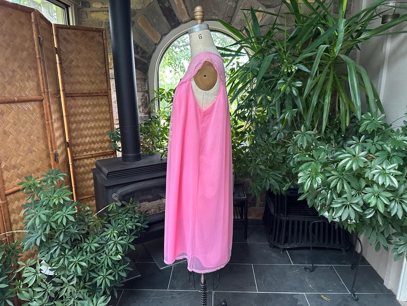 Puede incluir: Un camis&oacute;n rosa vintage en un maniqu&iacute;. El camis&oacute;n es de color rosa vibrante con detalles de encaje alrededor del escote y el dobladillo. La tela parece ligera y fluida. El maniqu&iacute; se exhibe en una habitaci&oacute;n con plantas y una chimenea.