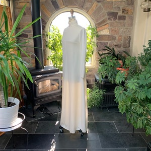 Peut inclure: Une longue robe blanche &agrave; col montant et manches longues est pr&eacute;sent&eacute;e sur un mannequin. La robe est longue et semble &ecirc;tre faite d'un tissu doux et fluide. Le mannequin est plac&eacute; dans une v&eacute;randa avec des plantes et un mur en pierre.