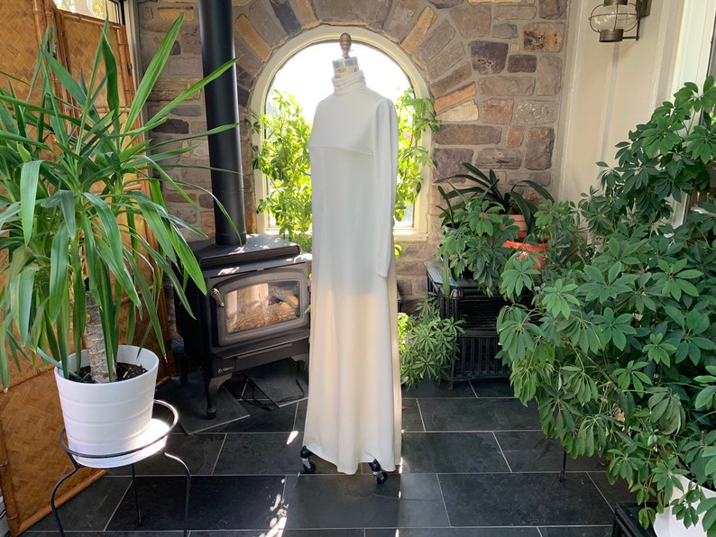 Peut inclure: Une longue robe blanche &agrave; col montant et manches longues est pr&eacute;sent&eacute;e sur un mannequin. La robe est longue et semble &ecirc;tre faite d'un tissu doux et fluide. Le mannequin est plac&eacute; dans une v&eacute;randa avec des plantes et un mur en pierre.