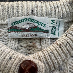 K&ouml;nnte beinhalten: Nahaufnahme eines cremefarbenen Aran-Pullovers mit strukturiertem Strickmuster. Der Pullover hat einen braunen Knopf und ein Etikett mit der Aufschrift "Aran Crafts Ireland 100% Wool M". Der Pullover wird in Irland hergestellt.