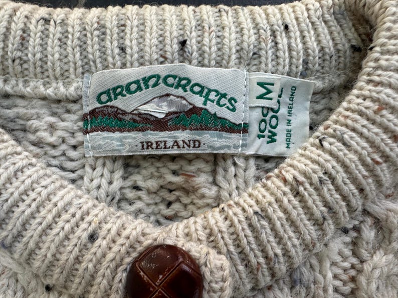 K&ouml;nnte beinhalten: Nahaufnahme eines cremefarbenen Aran-Pullovers mit strukturiertem Strickmuster. Der Pullover hat einen braunen Knopf und ein Etikett mit der Aufschrift "Aran Crafts Ireland 100% Wool M". Der Pullover wird in Irland hergestellt.