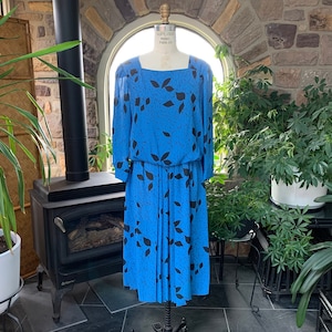 May include: A vintage blue dress with a black leaf pattern. The dress features a square neckline, long sleeves, and a pleated skirt. The dress is displayed on a mannequin in front of a window.