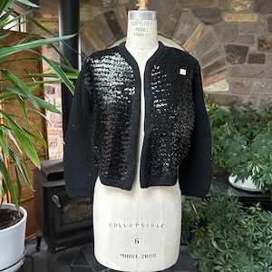 May include: Black cropped cardigan with sequin embellishments on the front panels, displayed on a vintage dress form. The open-front sweater has a round neckline and long sleeves. The dress form is labeled "Collapsible Model 2008".