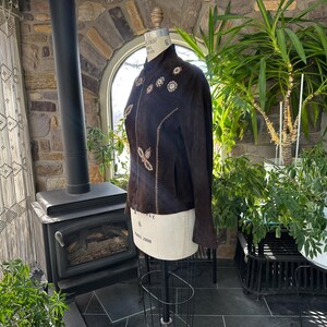 May include: A dark brown suede jacket with floral embroidery and a mandarin collar. The jacket is displayed on a mannequin, showcasing its tailored fit and decorative details. The jacket's design features a series of embroidered flowers.