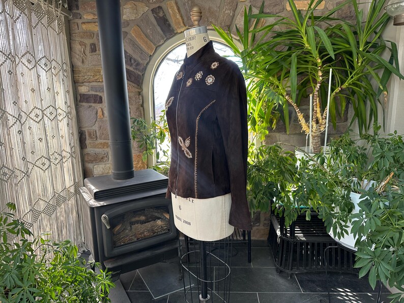 May include: A dark brown suede jacket with floral embroidery and a mandarin collar. The jacket is displayed on a mannequin, showcasing its tailored fit and decorative details. The jacket's design features a series of embroidered flowers.