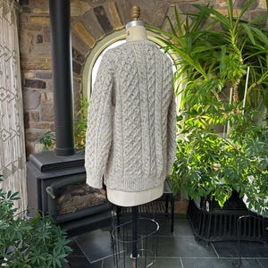 K&ouml;nnte beinhalten: Ein hellbeige Zopfmuster-Pullover, der auf einer Schaufensterpuppe pr&auml;sentiert wird. Der Pullover hat ein strukturiertes Muster und lange &Auml;rmel. Der Hintergrund umfasst einen Kamin, Pflanzen und einen dekorativen Vorhang, was eine gem&uuml;tliche, rustikale &Auml;sthetik schafft.