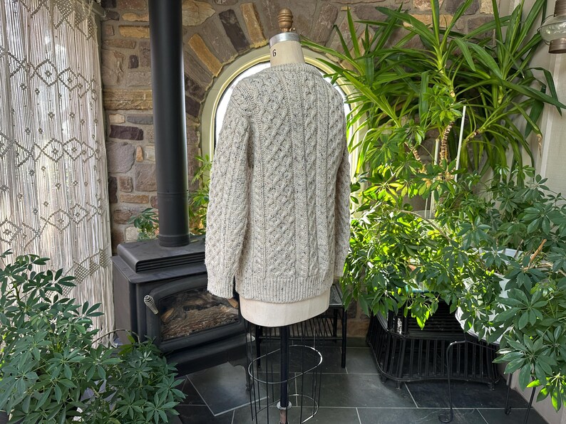 K&ouml;nnte beinhalten: Ein hellbeige Zopfmuster-Pullover, der auf einer Schaufensterpuppe pr&auml;sentiert wird. Der Pullover hat ein strukturiertes Muster und lange &Auml;rmel. Der Hintergrund umfasst einen Kamin, Pflanzen und einen dekorativen Vorhang, was eine gem&uuml;tliche, rustikale &Auml;sthetik schafft.