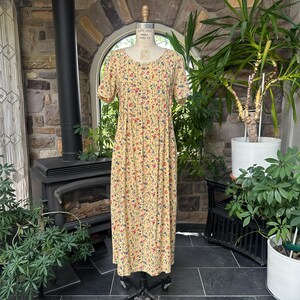 Könnte beinhalten: Ein gelbes Vintage-Kleid mit Blumenmuster, kurzen Ärmeln und rundem Ausschnitt. Das Kleid hat ein Muster aus kleinen roten, blauen und gelben Blumen. Das Kleid wird auf einer Schaufensterpuppe vor einer Steinmauer und Pflanzen präsentiert.