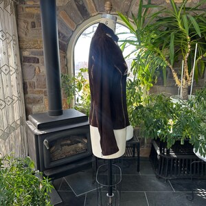 May include: A dark brown suede jacket displayed on a mannequin. The jacket features decorative stitching along the seams and a high collar. The mannequin is on a metal stand, and the background includes a stone wall, plants, and a fireplace.