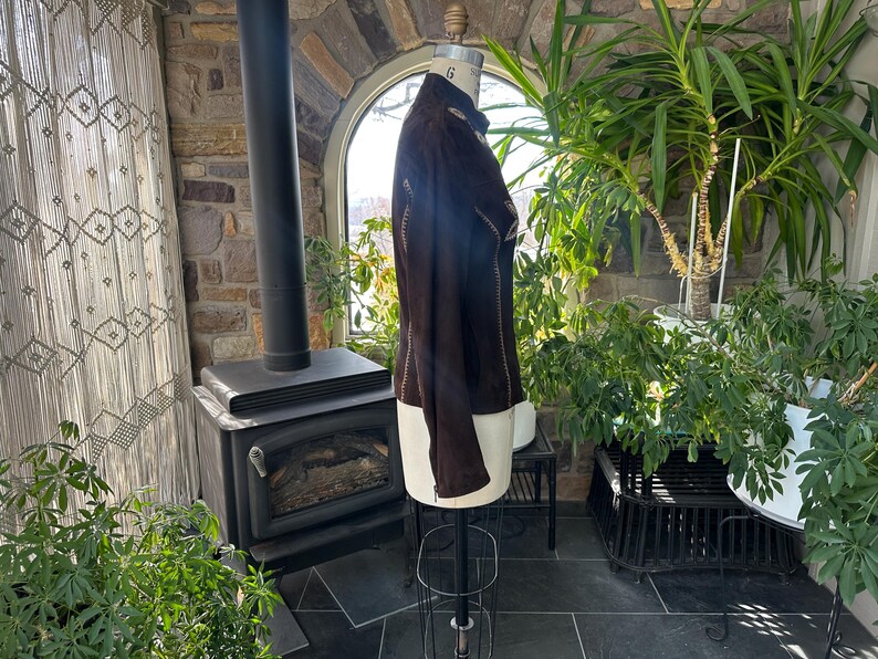 May include: A dark brown suede jacket displayed on a mannequin. The jacket features decorative stitching along the seams and a high collar. The mannequin is on a metal stand, and the background includes a stone wall, plants, and a fireplace.