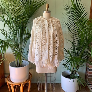 Op de afbeelding: Crèmekleurige, open geweven gehaakte cape met franjes. De cape heeft een knoopsluiting aan de voorkant en een decoratief, open patroon. Het kledingstuk wordt op een mannequin gepresenteerd, waarbij het ontwerp en de textuur worden getoond. Vintage stijl.