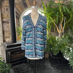 May include: A long-sleeved cardigan in shades of teal, blue, and purple, displayed on a mannequin. The sweater features a button-down front and a striped pattern. The garment is made of a soft, possibly knitted material, and is suitable for layering.