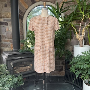 Puede incluir: Un vestido recto de manga corta con un patrón geométrico en color naranja claro y blanco. El vestido presenta un cuello redondo alto con un detalle de ribete rectangular en marrón oscuro y dos bolsillos de parche a juego en la parte delantera.