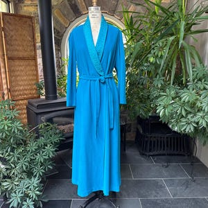 Könnte beinhalten: Ein langer, türkisfarbener Morgenmantel mit passendem Gürtel wird auf einer Schaufensterpuppe präsentiert. Der Morgenmantel hat einen strukturierten Kragen und Manschetten. Der Hintergrund umfasst einen Kamin, Pflanzen und einen dekorativen Paravent.