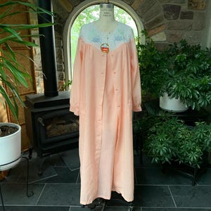 May include: A peach-colored, button-down robe with long sleeves, displayed on a mannequin. The robe features a white yoke with embroidered floral details. The garment is floor-length and appears to be made of a soft, lightweight fabric.
