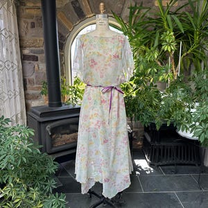 Può includere: Un abito vintage con stampa floreale su un manichino. L'abito presenta una sovrapposizione trasparente con maniche corte e una cintura di nastro viola. Lo sfondo include un muro di pietra, una stufa nera e una vegetazione lussureggiante.