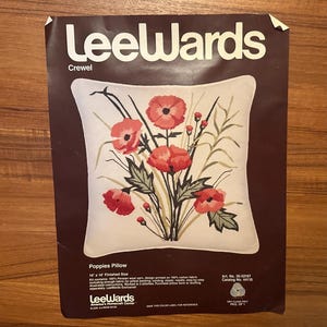May include: A Leewards Crewel Poppies Pillow kit package featuring a white square pillow with a red poppy floral design. The dark brown packaging includes the text Leewards, Poppies Pillow, and 14 x 14 Finished Size with kit details.