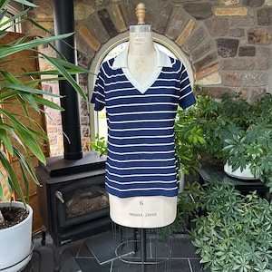 Peut inclure: Un t-shirt à manches courtes rayé bleu marine et blanc avec un col blanc et un col en V. Le t-shirt est présenté sur un mannequin. Le mannequin est sur un support en métal. L'arrière-plan comprend des plantes et un mur en pierre.