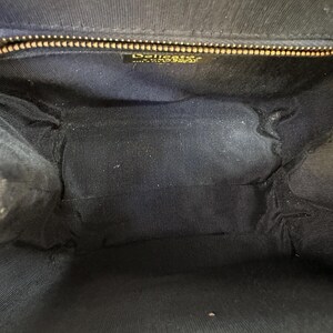 May include: The interior of a black handbag featuring a dark fabric lining. A gold-toned metal zipper runs across the top opening. A small rectangular label is attached below the zipper, displaying the text Delicato and Made in Italy.