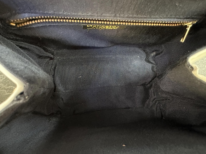 May include: The interior of a black handbag featuring a dark fabric lining. A gold-toned metal zipper runs across the top opening. A small rectangular label is attached below the zipper, displaying the text Delicato and Made in Italy.