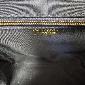 May include: The interior of a dark navy blue handbag featuring a gold-toned metal zipper closure at the top. A rectangular black fabric label is stitched inside, displaying the gold text Delicato by MARKAY MADE IN USA.