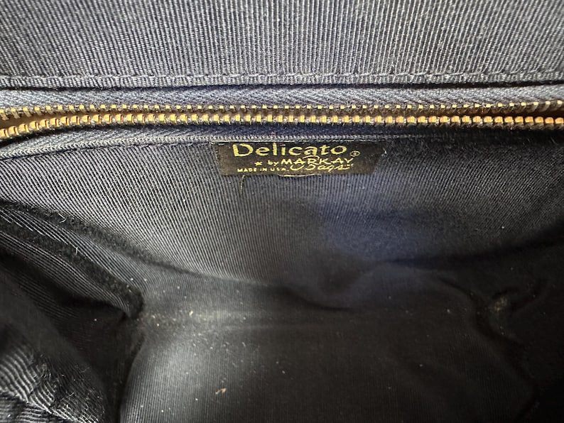 May include: The interior of a dark navy blue handbag featuring a gold-toned metal zipper closure at the top. A rectangular black fabric label is stitched inside, displaying the gold text Delicato by MARKAY MADE IN USA.