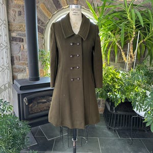 May include: Olive green vintage coat with a double-breasted design. The coat features a wide collar and decorative buttons. The coat is displayed on a mannequin in front of a fireplace and plants.