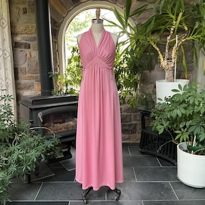 May include: A vintage pink halter-neck maxi dress. The dress features a fitted bodice with a pleated design and a flowing skirt. The dress is displayed on a mannequin in a room with plants and a fireplace.