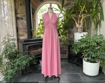 May include: A vintage pink halter-neck maxi dress. The dress features a fitted bodice with a pleated design and a flowing skirt. The dress is displayed on a mannequin in a room with plants and a fireplace.