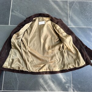 May include: A brown suede jacket with a light tan satin lining. The jacket is open, revealing the interior lining and the label "GEMETS". The jacket has a classic design with a collar and long sleeves.