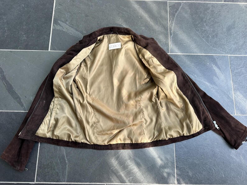 May include: A brown suede jacket with a light tan satin lining. The jacket is open, revealing the interior lining and the label "GEMETS". The jacket has a classic design with a collar and long sleeves.