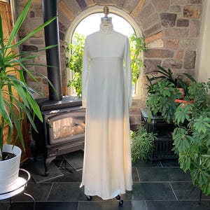 Peut inclure: Longue robe ivoire &agrave; manches longues et col montant. La robe est pr&eacute;sent&eacute;e sur un mannequin, mettant en valeur sa silhouette fluide. La robe semble &ecirc;tre faite d'un tissu l&eacute;ger, adapt&eacute; aux occasions formelles.