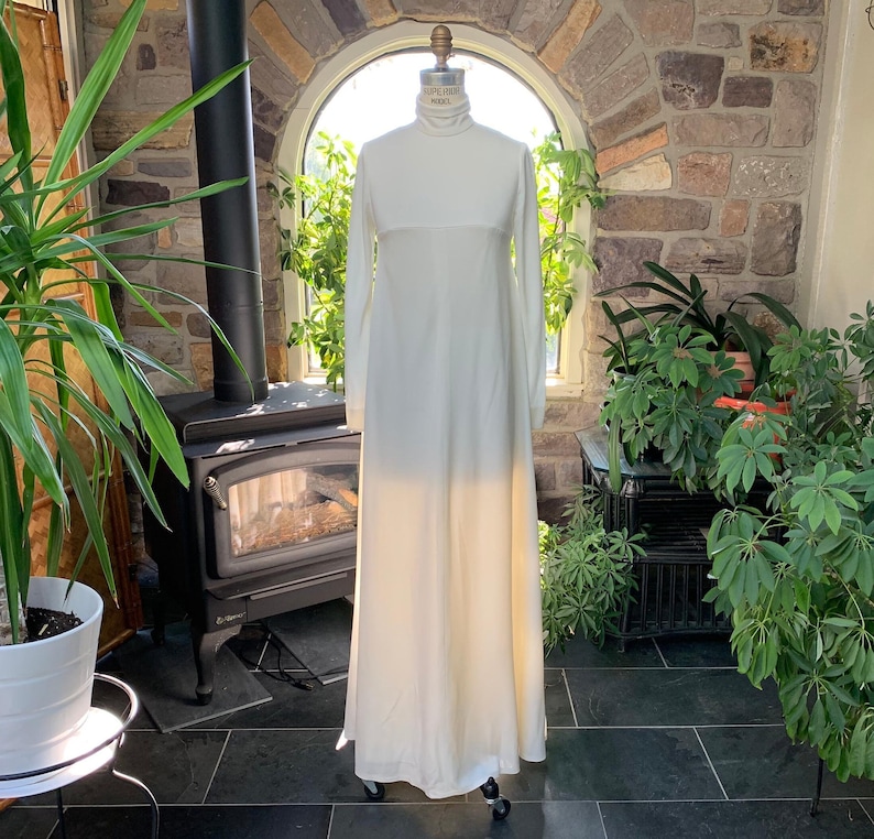 Peut inclure: Longue robe ivoire &agrave; manches longues et col montant. La robe est pr&eacute;sent&eacute;e sur un mannequin, mettant en valeur sa silhouette fluide. La robe semble &ecirc;tre faite d'un tissu l&eacute;ger, adapt&eacute; aux occasions formelles.