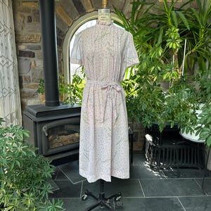 May include: A light pink vintage dress with short sleeves and a matching belt. The dress features a delicate floral pattern and is displayed on a dress form. The background includes a fireplace, plants, and a window.