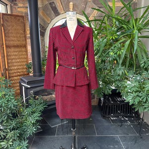 May include: A red and black houndstooth pattern skirt suit displayed on a mannequin. The suit includes a blazer with a black collar and a matching skirt. A thin black belt cinches the waist. The suit is displayed in a room with plants and a fireplace.