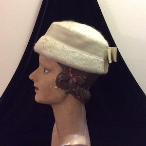 May include: A vintage cream-colored hat with a rounded crown and a band around the base. The hat features a fluffy, textured brim and a small bow detail on the side. The hat is displayed on a mannequin head.