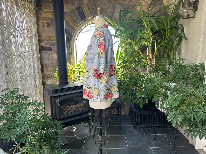 Może przedstawiać: Jasnoniebieska koszula z tropikalnym wzorem kwiatowym z czerwonych kwiat&oacute;w i ż&oacute;łtych ananas&oacute;w. Koszula jest prezentowana na manekinie. Tło obejmuje rośliny, kominek i okno.