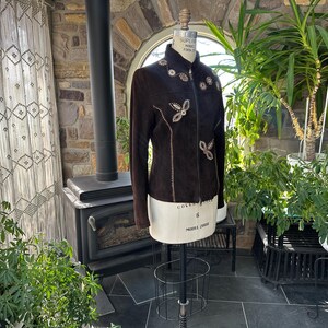 May include: A dark brown suede jacket with decorative floral and butterfly-shaped embellishments. The jacket is displayed on a mannequin, showcasing its tailored fit and intricate detailing. The jacket has a high collar and long sleeves.