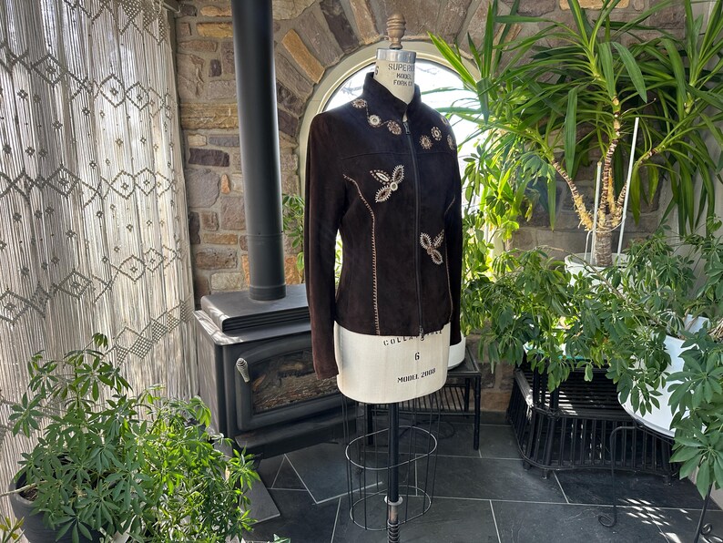 May include: A dark brown suede jacket with decorative floral and butterfly-shaped embellishments. The jacket is displayed on a mannequin, showcasing its tailored fit and intricate detailing. The jacket has a high collar and long sleeves.