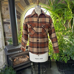 May include: A long-sleeve button-up shirt featuring a plaid pattern in shades of tan, brown, and rust orange. The shirt includes a pointed collar, two chest pockets with buttoned flaps, and a front button closure, displayed on a dress form.