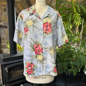 Może przedstawiać: Jasnoniebieska koszula zapinana na guziki z kr&oacute;tkim rękawem z tropikalnym nadrukiem kwiatowym. Koszula ma czerwone kwiaty hibiskusa, ż&oacute;łte ananasy i zielone liście. Koszula jest prezentowana na manekinie.