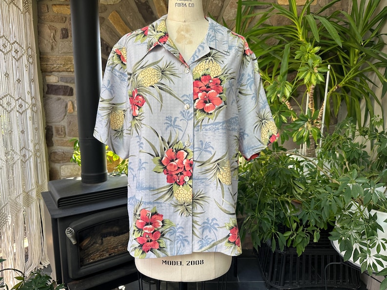 Może przedstawiać: Jasnoniebieska koszula zapinana na guziki z kr&oacute;tkim rękawem z tropikalnym nadrukiem kwiatowym. Koszula ma czerwone kwiaty hibiskusa, ż&oacute;łte ananasy i zielone liście. Koszula jest prezentowana na manekinie.