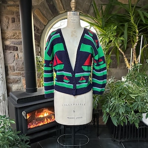 May include: A navy blue, green, and red knit cardigan sweater featuring a repeating sailboat pattern. The garment has a V-neckline, long sleeves, and a front button closure. It is displayed on a beige dress form with visible text markings.