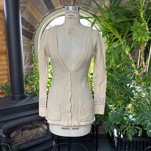 Puede incluir: Cárdigan color crema con cuello en V y cierre de botones, exhibido en un maniquí. Las mangas largas tienen puños a rayas en colores pastel. El suéter tiene dos bolsillos delanteros y un diseño de punto texturizado.