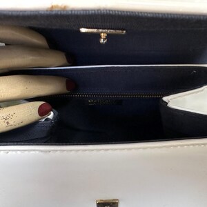 May include: A white patent leather handbag with a structured rectangular shape and a glossy finish. The interior is lined with dark blue fabric and features a gold-toned metal clasp closure at the top. A small rectangular label reads Delicato.