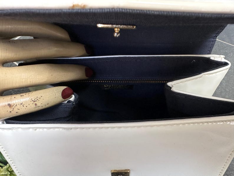 May include: A white patent leather handbag with a structured rectangular shape and a glossy finish. The interior is lined with dark blue fabric and features a gold-toned metal clasp closure at the top. A small rectangular label reads Delicato.