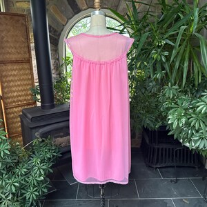 Puede incluir: Un camis&oacute;n rosa vintage en un maniqu&iacute;. El camis&oacute;n es de un rosa vibrante liso con un canes&uacute; transparente y mangas cortas. El escote y el dobladillo est&aacute;n adornados con delicado encaje. El fondo incluye vegetaci&oacute;n y una chimenea.