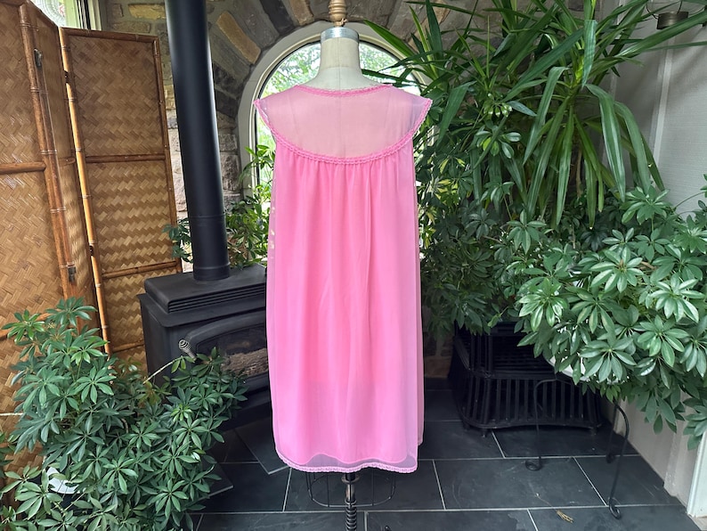 Puede incluir: Un camis&oacute;n rosa vintage en un maniqu&iacute;. El camis&oacute;n es de un rosa vibrante liso con un canes&uacute; transparente y mangas cortas. El escote y el dobladillo est&aacute;n adornados con delicado encaje. El fondo incluye vegetaci&oacute;n y una chimenea.